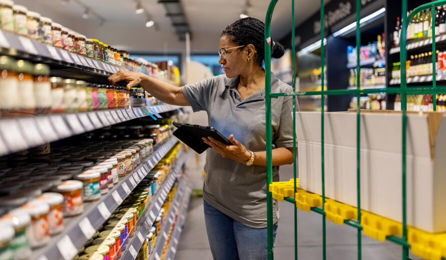 Elderly female supermarket worker managing grocery inventory with a digital tablet.