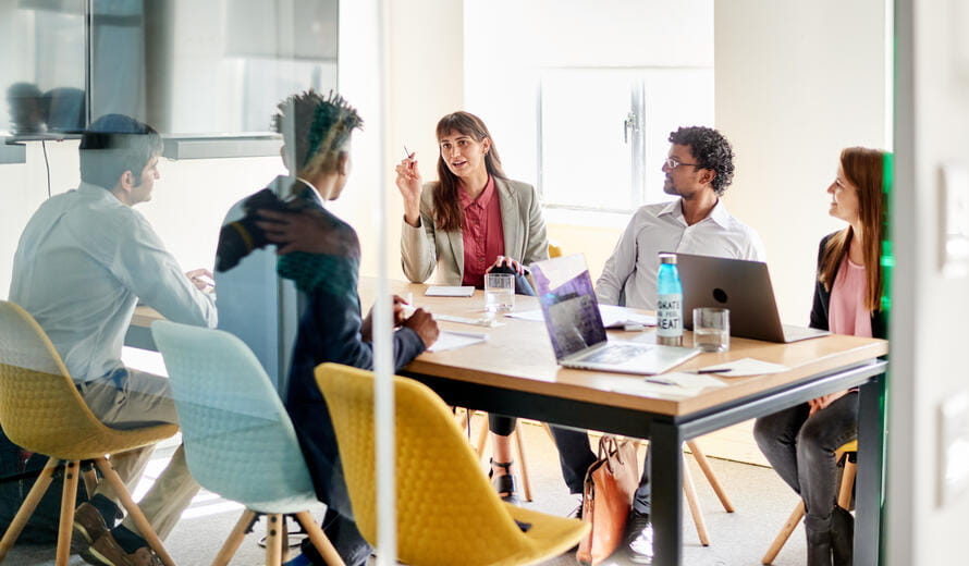 Group of diverse young businesspeople discussing work together while sitting around a table inside of an office boardroom
