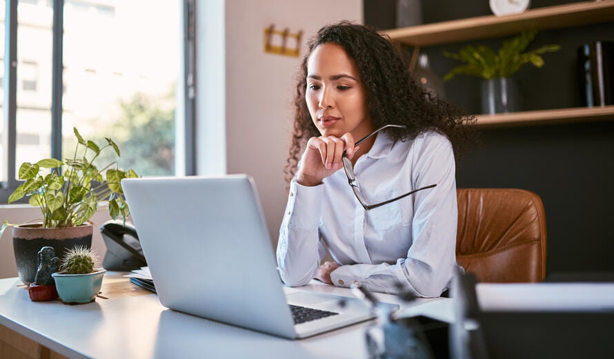 Young and ambitious mixed race entrepreneur sitting alone and thinking while browsing the internet on a laptop in an office. Hispanic business woman using a computer to plan her freelancing startup