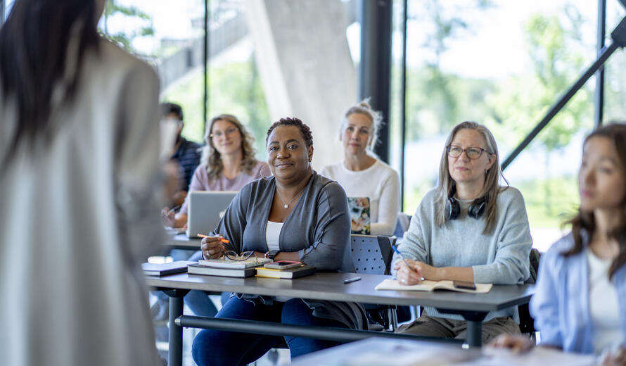 A large group of diverse mature students, sit together in rows as they listen attentively to their teacher explain the lesson. They are each dressed casually and have notebooks, textbooks, laptops and papers out in front of them for reference and taking notes.
