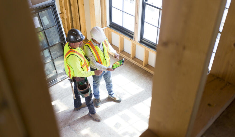 A contract supervisor talks with a contractor while in a room a room of a residential building and wearing a hard hat, safety vest, PPE and using a mobile device