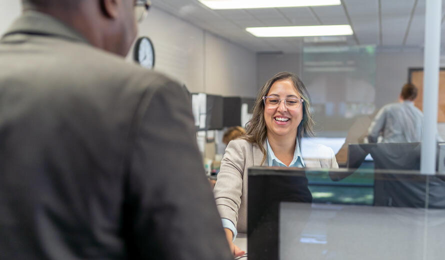 A female customer service representative of Latin descent smiles while greeting an unrecognizable male black customer at the reception desk of a medical clinic or bank.