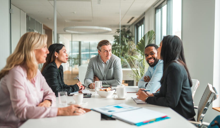 Multi-ethnic team of mid adult male and female managers sitting in a meeting room, leaning on the table and discussing. Waist up image, front view of the Caucasian and African man, side view of the Caucasian and Asian women.