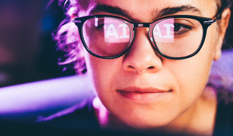 A person wearing glasses with a reflection of artificial intelligence technology on their laptop.