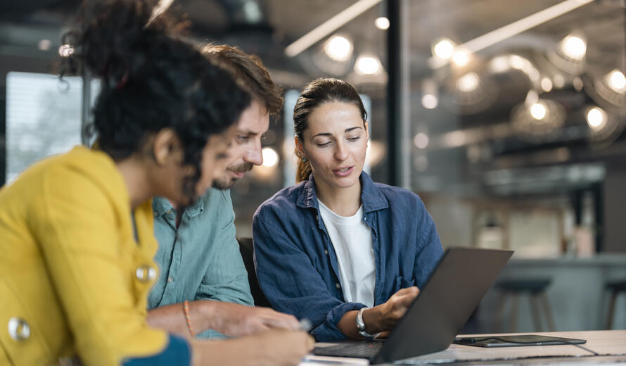 Small group of co-workers working together in a startup business office space. A team of young people in a modern office is discussing their project. Group of business people working. The team are sitting at a board room table looking at a laptop computer.