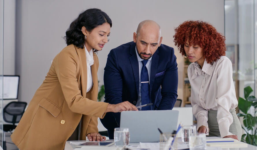 Three diverse business colleagues lean over a laptop reviewing documents.