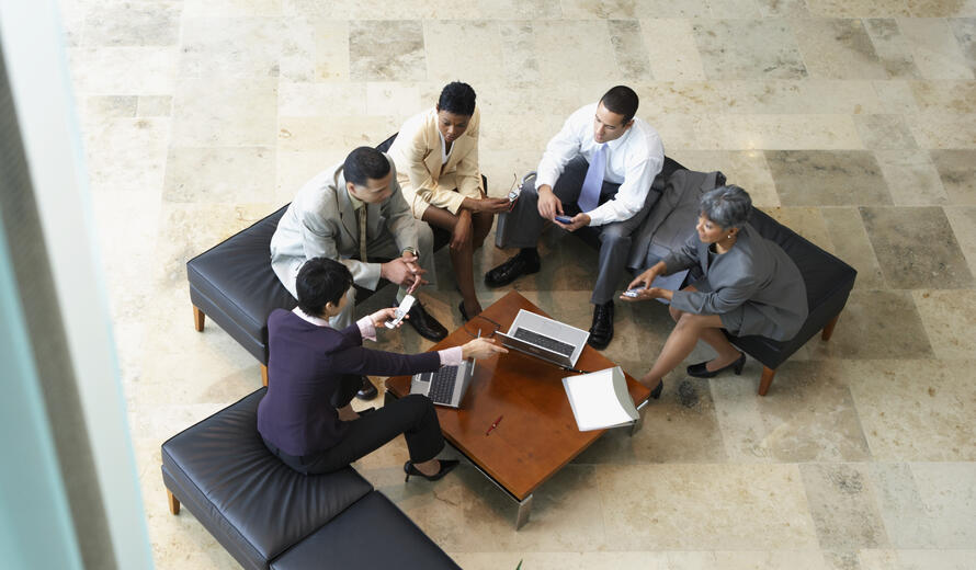 An overhead image of a group of professionals sitting in an office lounge area, collaborating around a table with laptops and documents.