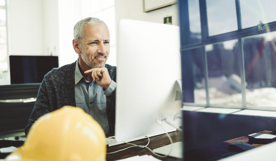 A mature architect smiling while using a computer at his desk in a brightly lit office