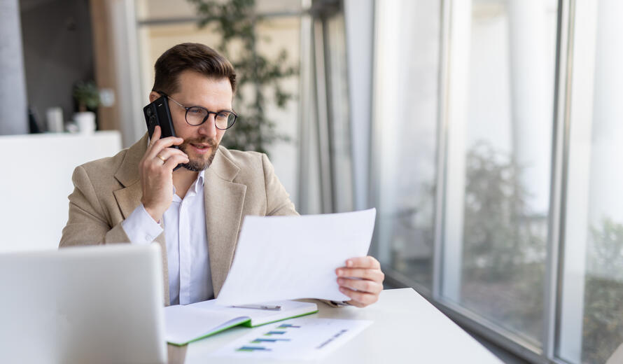 Engaged man is reviewing documents while on a phone call in a bright modern office, reflecting a mix of professionalism and effective communication.
