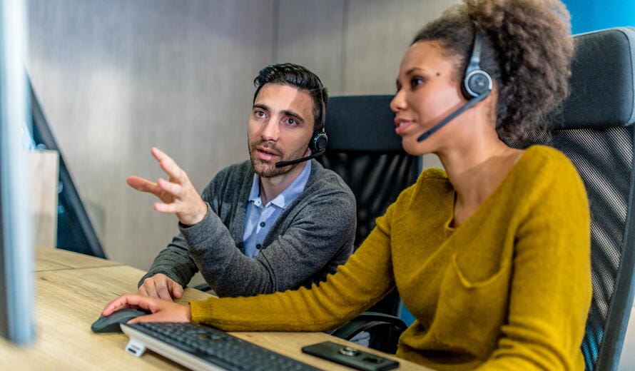 Photo of a man working in a call centre helping his female colleague while they are using a computer.