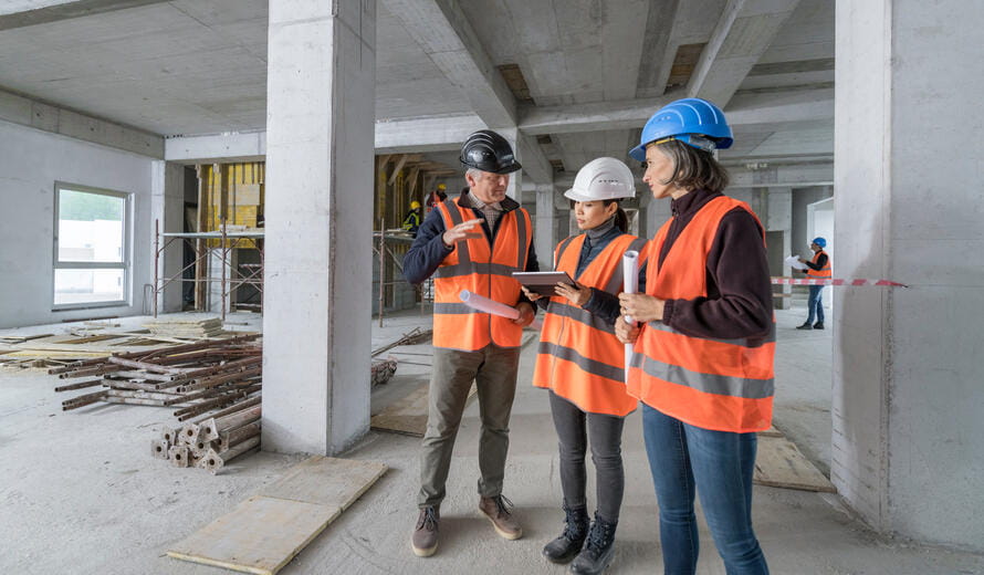 individuals wearing safety helmets and high-visibility vests reviewing construction plans using a tablet and printed drawings inside a partially completed concrete building structure with scaffolding and materials visible.