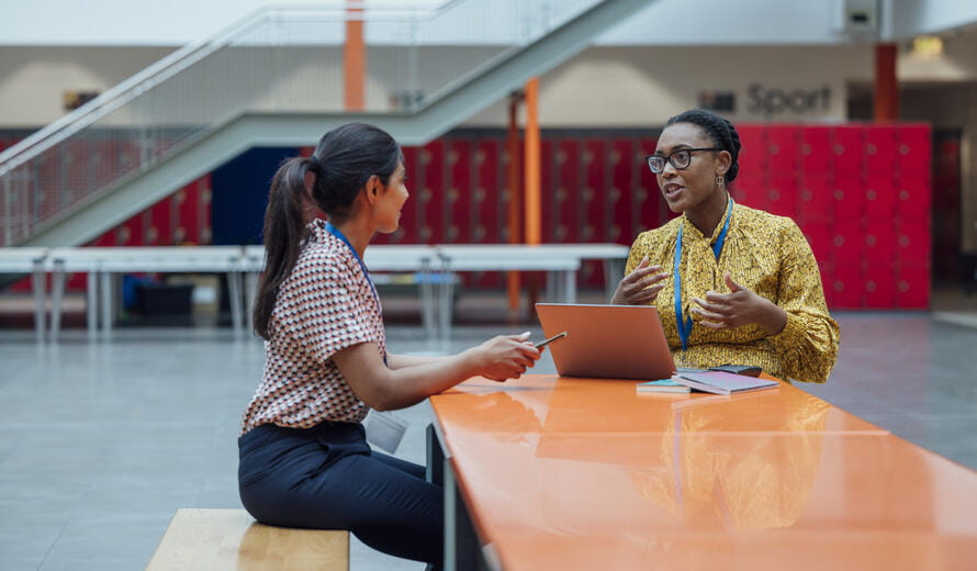 Two female teachers sitting at a table, having a discussion in the lobby area in the school they work at in Gateshead, North East England.