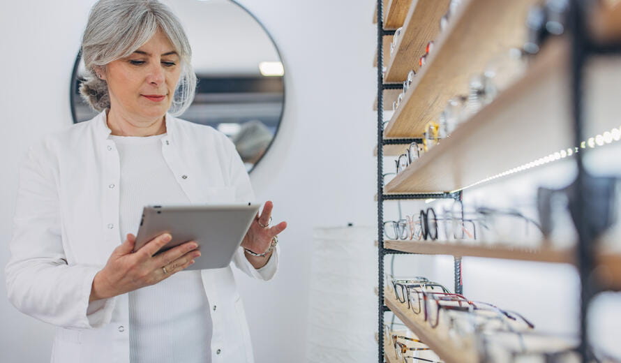 An older woman in a white coat, possibly an employee, uses a tablet to check the eyewear display in an optical store, ensuring the arrangement and quality of the glasses.
