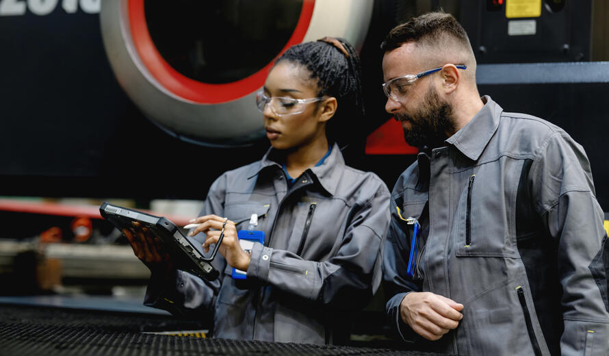 female engineer and supervisor with a mobile computer are working on engineering parts production line. They are discussing while standing by an automated CNC machine.