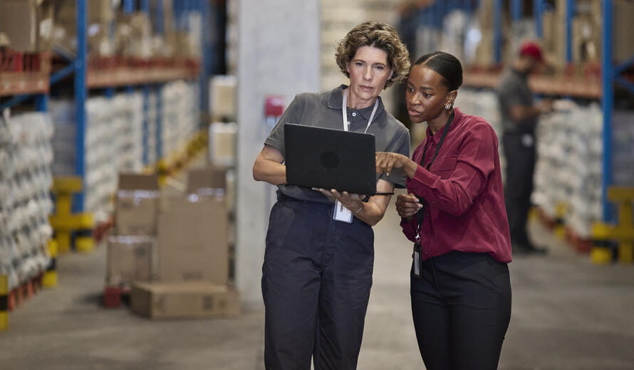 A female manager and an employee standing in a distribution warehouse, reviewing information together on a laptop among shelves stocked with inventory.