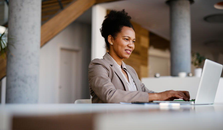 Smiling businesswoman sitting at a modern desk, working on a laptop in a stylish office, engaging in online communication and managing tasks in a dynamic corporate environment