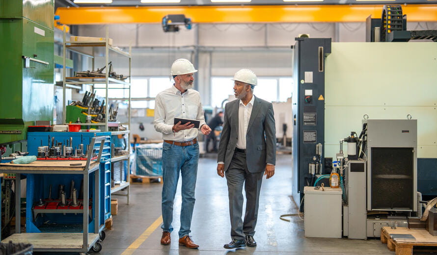 Mature Hispanic manager collaborating with Caucasian engineer in a factory setting featuring industrial machinery. Both professionals wear safety helmets and formal attire, engaging in a focused discussion while reviewing data on a digital tablet