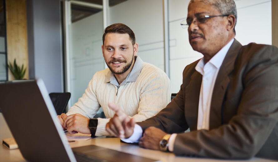 A mature Black businessman points at a laptop screen, guiding a younger white male colleague during a discussion or collaboration session at an office meeting table.