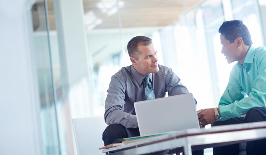 Two young businessmen working together while sharing a laptop between them