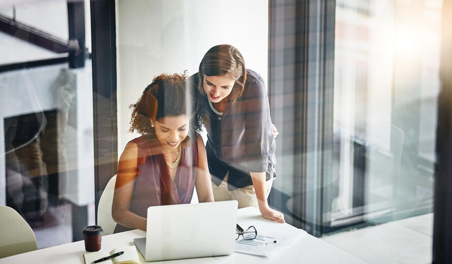 Two business women collaborating at a laptop in a modern office setting, reviewing information together while working at a desk.